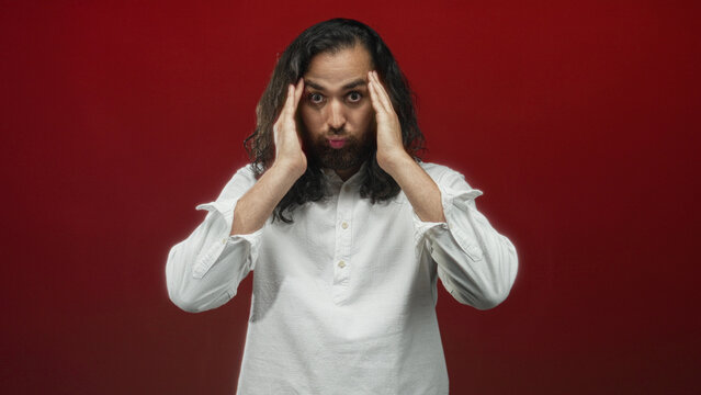 Young hispanic arab man with beard pressing temples with both hands, eyes closed, wearing white shirt in red studio; stress introspection headache.