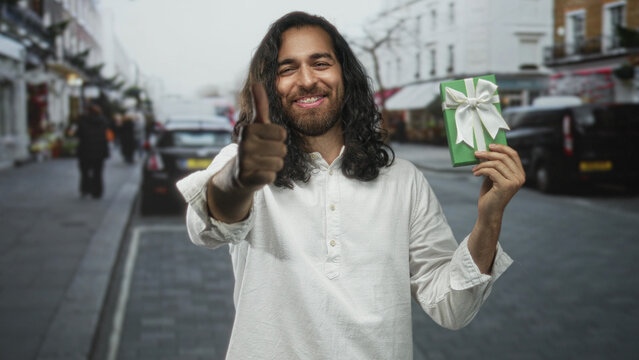 Man smiling, thumbs up with right hand while holding a green wrapped gift with white bow on a city street market; joy generosity giving.