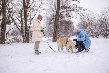 Elderly woman and her adult daughter spending time outdoors with their Golden Retriever on a snowy winter day. The daughter crouches to interact with the dog while the mother holds the leash