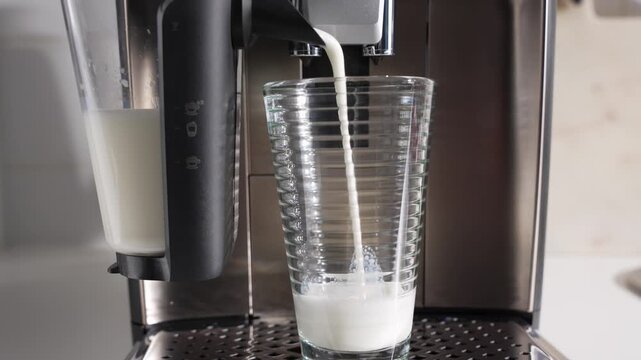 A tall glass fills with milk foam while espresso pours in. Automatic milk carafe visible.