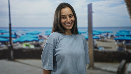 Woman smiling beside turquoise beach umbrellas on a studio set, wearing loose blue tshirt and relaxed posture with closed eyes; carefree joy.