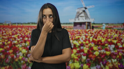 Young hispanic woman in black t shirt pinches nose while crossing arms among colorful tulip flowers near windmill building under blue sky; nuisance odor disgust.