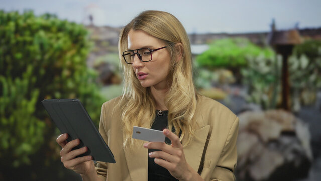 Woman outdoors in park using tablet and card, young blonde caucasian female in glasses focused on screen, surrounded by greenery suggesting online shopping or mobile banking.