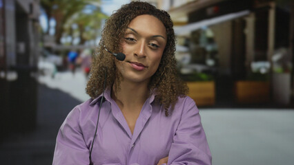 Transgender woman wearing a purple shirt and headset mic with arms crossed on a sunlit street;...