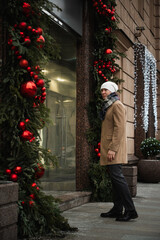 Adult caucasian man enters to luxury store for shopping. Xmas holiday. Man checking phone by festive Christmas decorations.