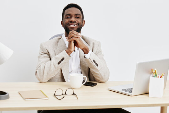 Confident smiling man in business suit sitting at desk with laptop, coffee cup, and glasses in bright modern office environment. People lifestyle concept, professional, positive mood, workplace