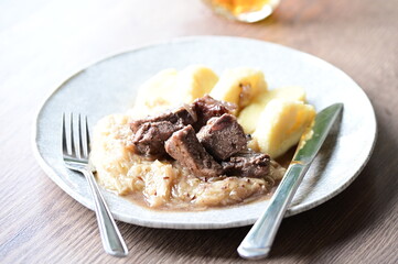 Traditional Czech dish of roasted wild boar with sauerkraut and dumplings, served on a plate in a cozy home-style setting. A hearty and flavorful meal representing authentic Czech cuisine and rustic c