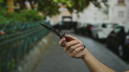 Man holding knife on city street background with blurred cars and greenery, emphasizing male hand in outdoor urban setting.