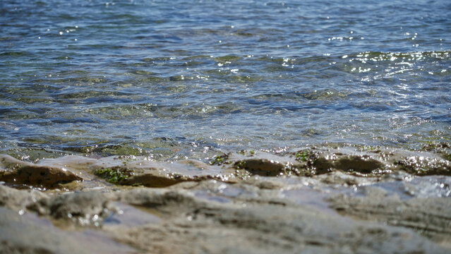 Mediterranean beach with clear sea under sunny sky, showing serene outdoor coastal scene with gentle waves and rocky shoreline sparkling in the bright sunlight.