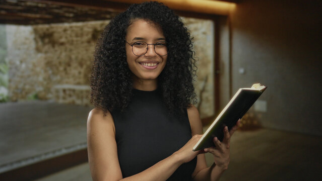 Woman reading book indoors with stone wall in background, wearing glasses and smiling, creating a cozy academic atmosphere showcasing a young, hispanic woman engaged in literature.