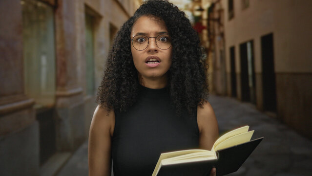 Young woman with glasses reading a book on an old town street, expressing curiosity and contemplation against a historic outdoor background.