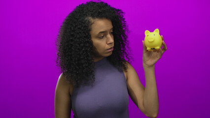 Woman holding yellow piggybank examines savings against pink background suggesting careful financial planning and thoughtful budgeting