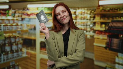 Woman holding canadian passport smiling confidently in supermarket background showing travel identification highlighting adult female traveler with red hair indoors