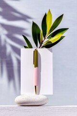 A stylish pen with green and pink accents is displayed on a smooth white stone, with a green leafy branch and palm leaf shadows in the background.