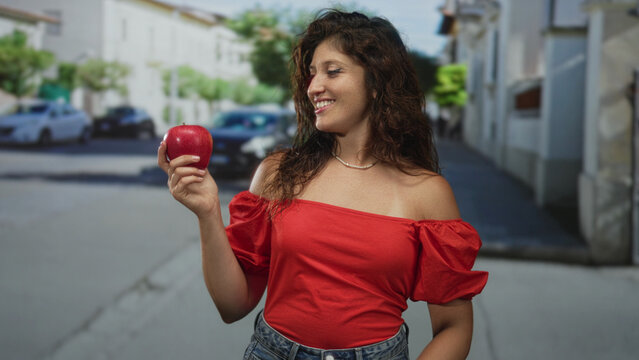 Young hispanic woman holding a red apple and smiling while waving on a city street wearing an off shoulder red top; joy healthy snack.