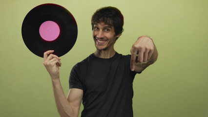 Man holds black vinyl record with pink label and points finger to it in green studio; music promotion excitement.