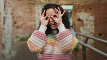 Young hispanic woman playfully posing with binocular fingers in front of ancient roman ruins on a...