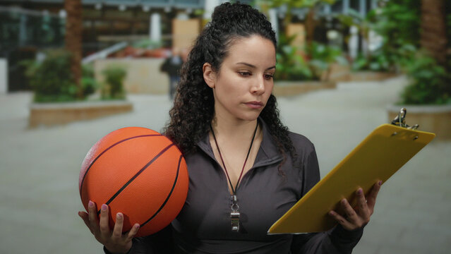 Woman with basketball outdoors on street holding clipboard looking at notes, wearing black top with whistle, surrounded by greenery in urban setting.
