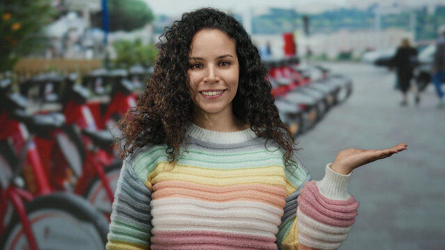 Young hispanic woman with curly hair extends hand in front of urban street with row of bicycles creating a friendly and welcoming atmosphere outdoors.