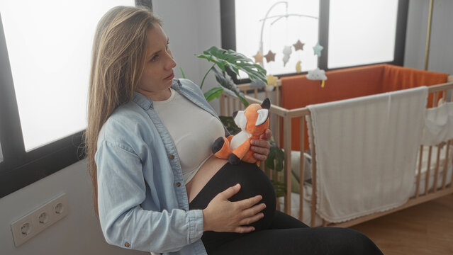 Pregnant woman sits in a serene baby room holding a toy fox gently on her belly, surrounded by a crib, soft lighting, and playful decor, embodying maternal warmth and anticipation. - Powered by Adobe