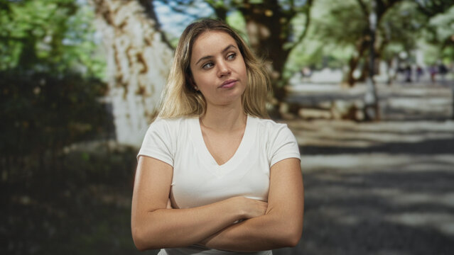 Young blonde woman arms crossed standing in forest park wearing white t shirt and skeptical expression; annoyance.