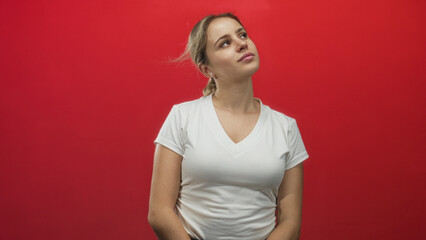 Young blonde woman tilting head and looking up in a red studio backdrop wearing a white v neck t...
