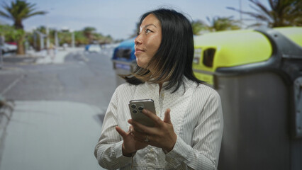 Woman tapping smartphone with hands on street by recycling container near parked cars and palm trees; sustainability calm.