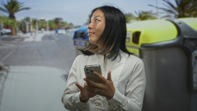Woman tapping smartphone with hands on street by recycling container near parked cars and palm trees; sustainability calm.