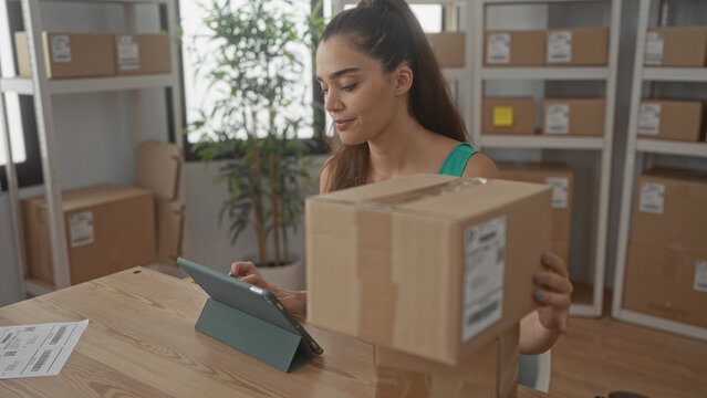 Woman holds cardboard package and checks shipping label at office building desk with stacked parcels and tablet; productivity organization.