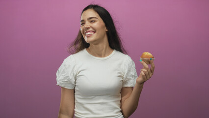 Woman holding a freshly baked muffin aloft in a pink studio setting showing a casual snack moment; joy wellness satisfaction.