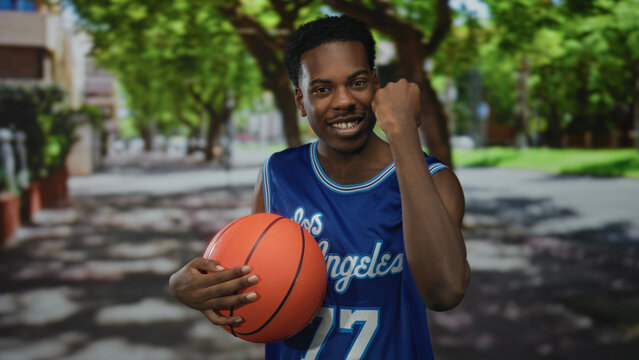 Man holds orange basketball and pumps fist on street under bright sunlight; exhilaration victory pride.