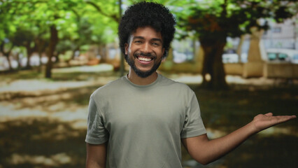 Fototapeta premium Man with afro hair smiling and presenting something with outstretched hand in a sunny park surrounded by lush green trees, embodying joy and enthusiasm outdoors.