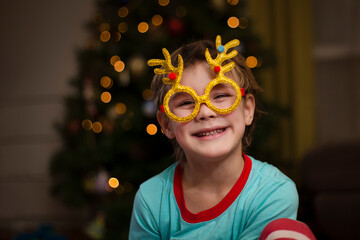 kid boy  Wearing carnival glasses with Christmas decor on near the Christmas tree at home. 