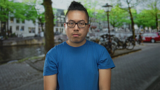Man in blue t shirt wearing glasses with eyes closed standing on a cobbled street by a canal with bicycles; introspective calm. - Powered by Adobe