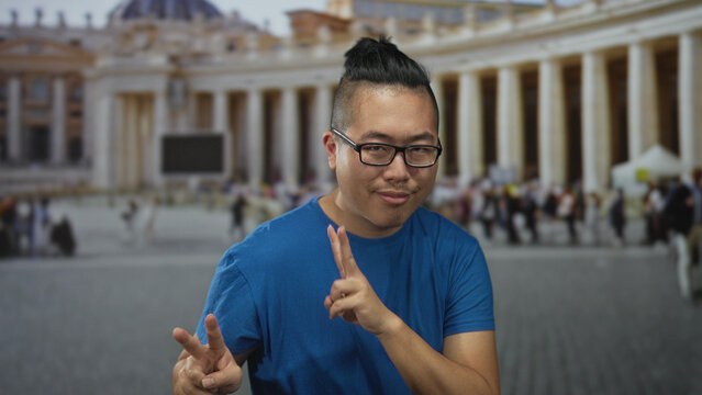 Man showing double peace sign with hands raised in st peter's square at the vatican outdoors, wearing blue t shirt and glasses; joy travel memory.