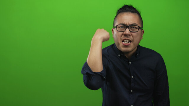 Chinese young man wearing glasses and a dark shirt clenches fist and grimaces in green studio; anger focus resolve. - Powered by Adobe
