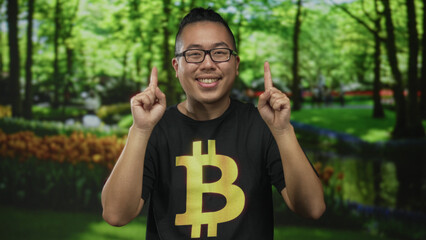 Young man wearing a black bitcoin t shirt smiles and points his index fingers upward in a sunlit forest park with green trees and lawn; crypto optimism.