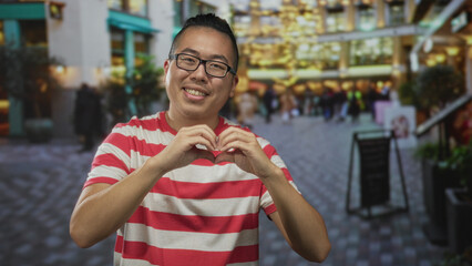 Young chinese man in red striped shirt and glasses forms heart with hands on crowded street...