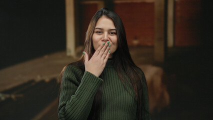 Woman sending kiss to camera stands in unfinished indoor construction site wearing green sweater,...