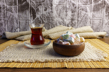 Traditional Turkish still life featuring a glass of black tea (çay) and a wooden bowl of colorful Turkish delight (lokum). Served on woven fabric and bamboo mat with a blurred stone background