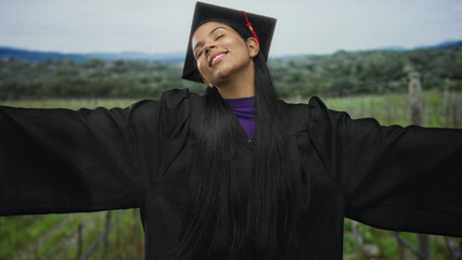 Naklejka premium Young latina woman in black graduation gown and mortarboard with red tassel spreads arms in green countryside field; academic achievement pride.