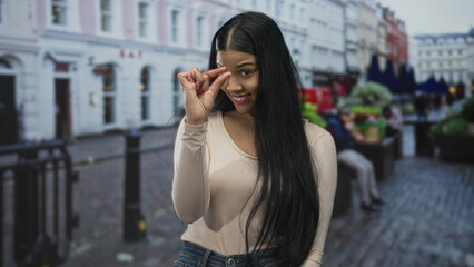 Fototapeta premium Young latin woman wearing beige top pinches fingers and smiles on street amid building facade under evening lights; teasing.