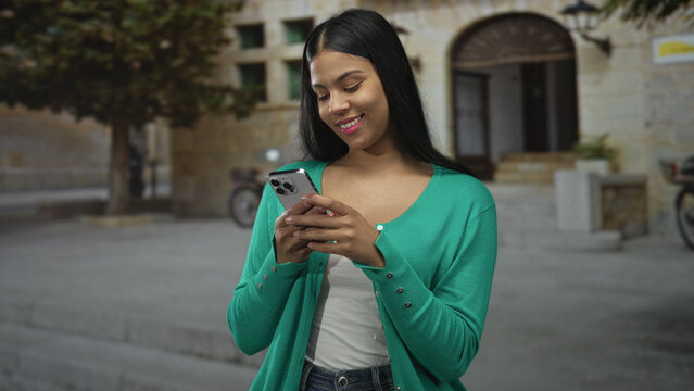 Young latin woman typing on smartphone on street while smiling and showing her hands with casual posture; social connection happiness.