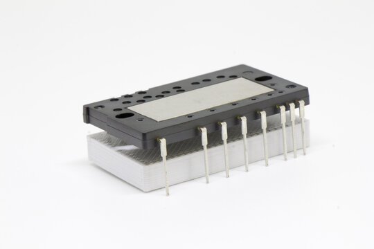 Angled close-up of a rectangular IPM (Intelligent Power Module) raised on a white spacer, showing dual rows of silver connector pins and a central metal heat-spreader plate, isolated on white