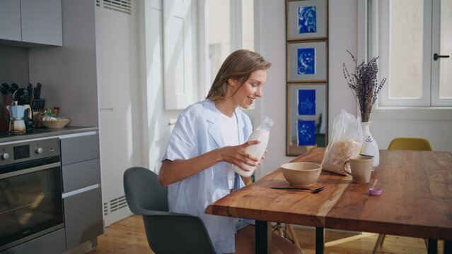 Breakfasting woman pouring milk into sweet cereal bowl. Relaxed lady sit table