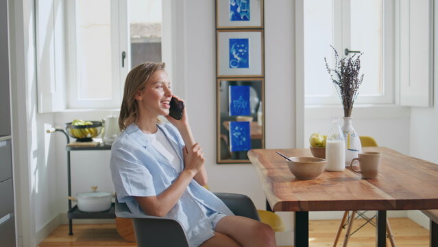 Relaxed woman talking smartphone at kitchen table. Smiling girl breakfasting