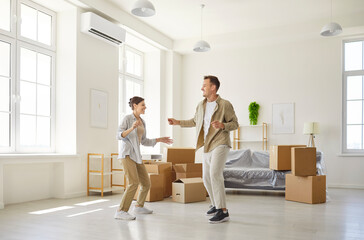 Young happy excited couple dancing amid moving boxes home. New apartment with tall windows and...