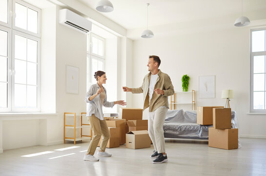 Young happy excited couple dancing amid moving boxes home. New apartment with tall windows and covered sofa shows relocation, move and rent. Idea celebration of new domestic beginnings.