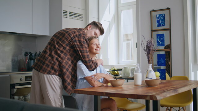 Happy couple hugging morning in sunny kitchen. Loving man kissing smiling woman