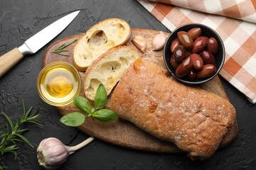 Fresh cut ciabatta, oil, olives, garlic and basil on dark textured table, flat lay